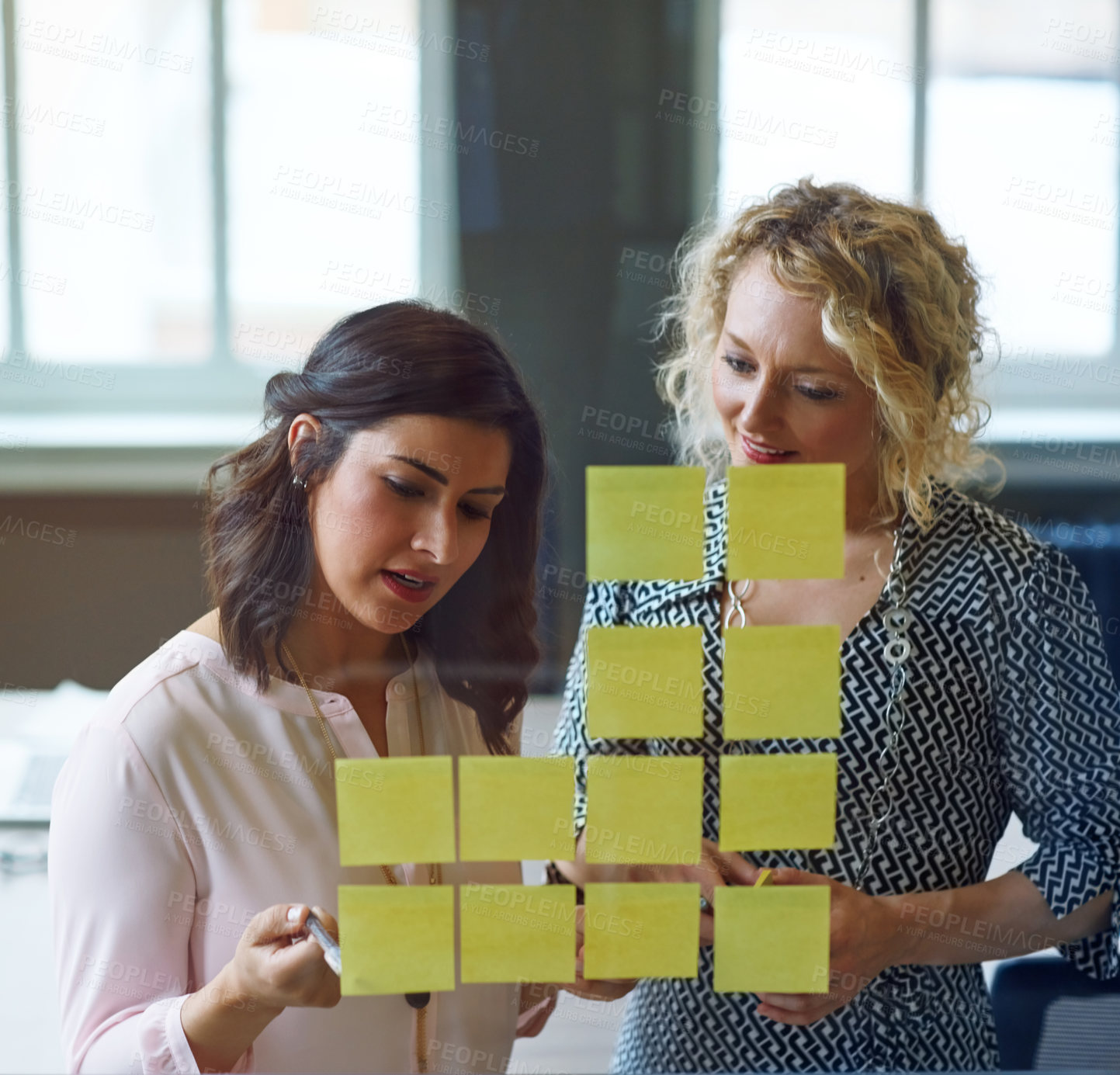 Buy stock photo Shot of two businesswomen brainstorming with adhesive notes on a glass wall in an office