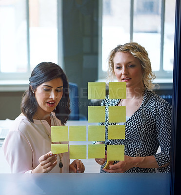 Buy stock photo Shot of two businesswomen brainstorming with adhesive notes on a glass wall in an office