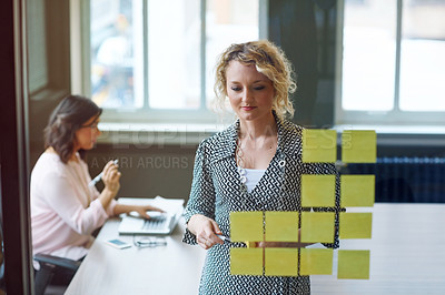 Buy stock photo Shot of a businesswoman reading adhesive notes on a glass wall with a colleague in the background