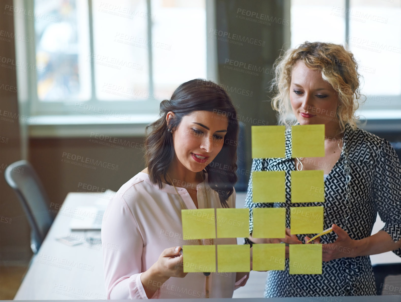 Buy stock photo Shot of two businesswomen brainstorming with adhesive notes on a glass wall in an office