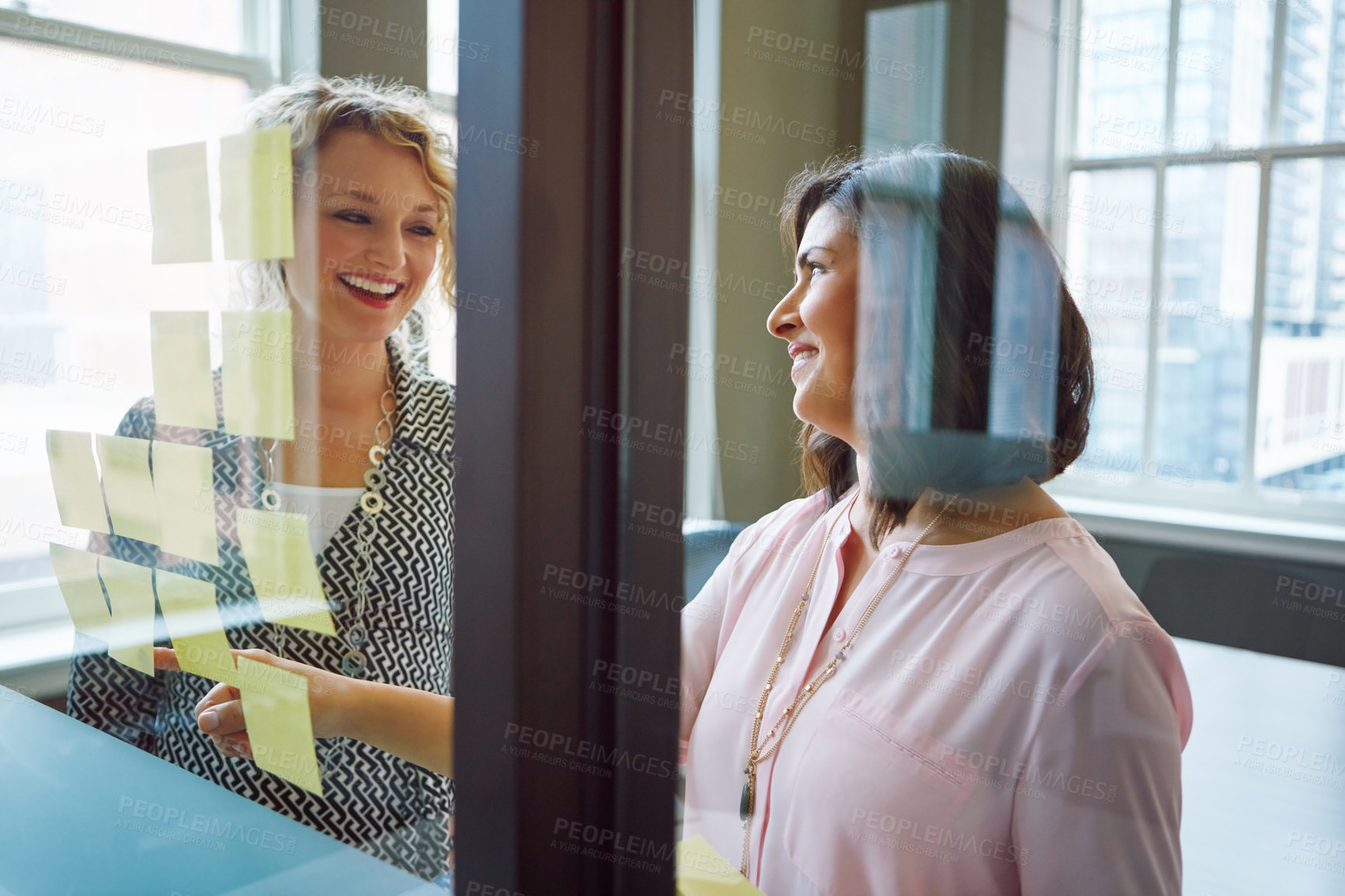 Buy stock photo Shot of two businesswomen brainstorming with adhesive notes on a glass wall in an office