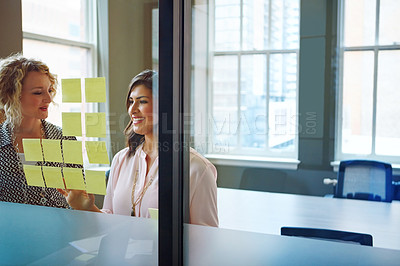 Buy stock photo Shot of two businesswomen brainstorming with adhesive notes on a glass wall in an office