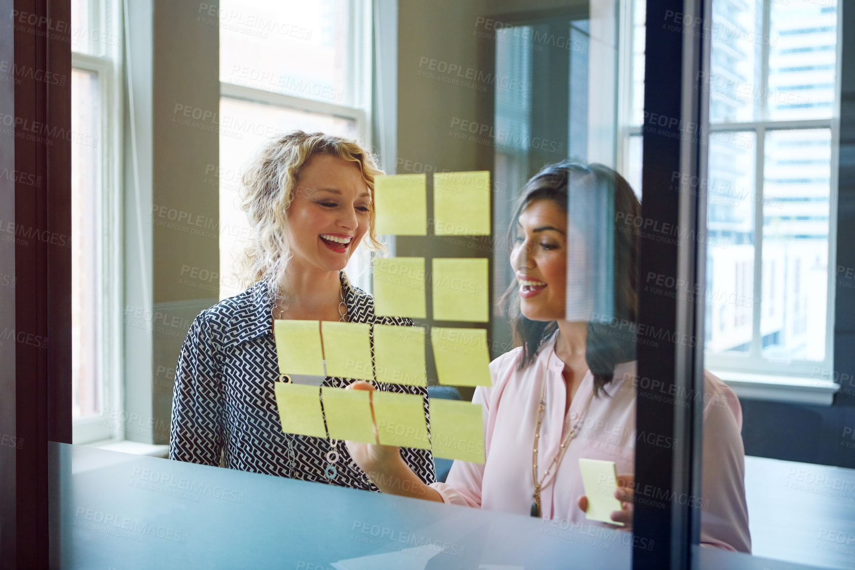 Buy stock photo Shot of two businesswomen brainstorming with adhesive notes on a glass wall in an office