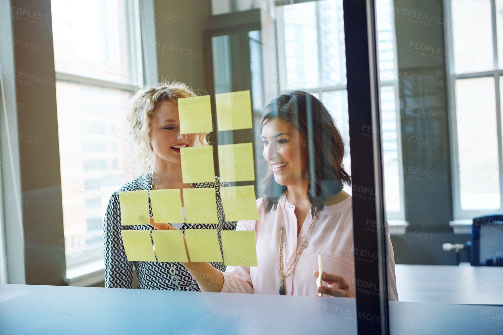 Buy stock photo Shot of two businesswomen brainstorming with adhesive notes on a glass wall in an office