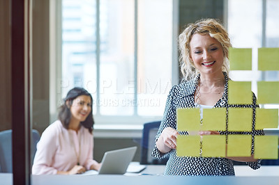 Buy stock photo Shot of a businesswoman reading adhesive notes on a glass wall with a colleague in the background