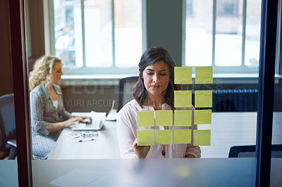 Buy stock photo Shot of a businesswoman reading adhesive notes on a glass wall with a colleague in the background