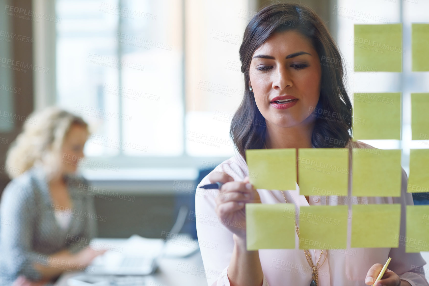 Buy stock photo Shot of a businesswoman reading adhesive notes on a glass wall with a colleague in the background