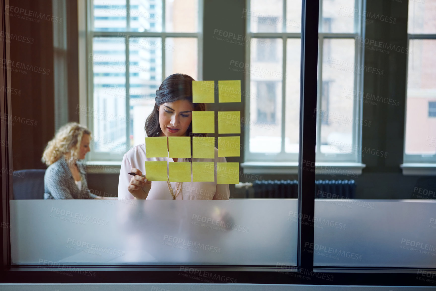 Buy stock photo Shot of a businesswoman reading adhesive notes on a glass wall with a colleague in the background