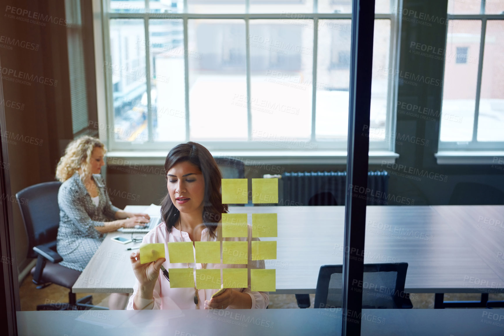 Buy stock photo Shot of a businesswoman reading adhesive notes on a glass wall with a colleague in the background