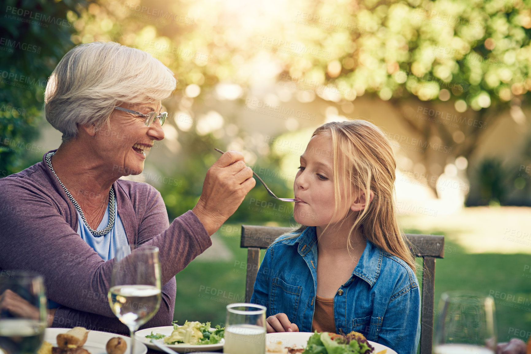 Buy stock photo Grandmother, girl and feeding at lunch, outdoor and happy with bonding, reunion and love in summer. People, grandma and child for food, helping hand or smile with care in backyard at family house