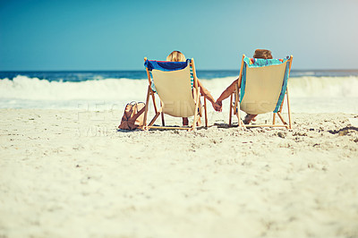 Buy stock photo Rearview shot of a young couple sitting hand in hand on loungers at the beach