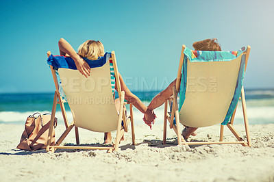 Buy stock photo Rearview shot of a young couple sitting hand in hand on loungers at the beach