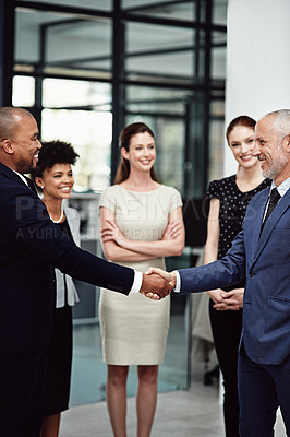 Buy stock photo Shot of two businessmen shaking hands while surrounded by their team at work