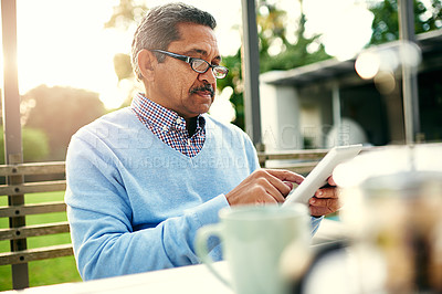 Buy stock photo Shot of an older man using a digital tablet while having his breakfast outdoors