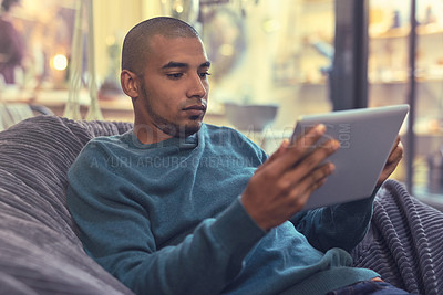 Buy stock photo Shot of a young man relaxing on a beanbag and using a digital tablet