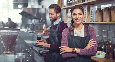 Buy stock photo Portrait of a confident young woman working in a cafe while her coworker operates a coffee machine in the background