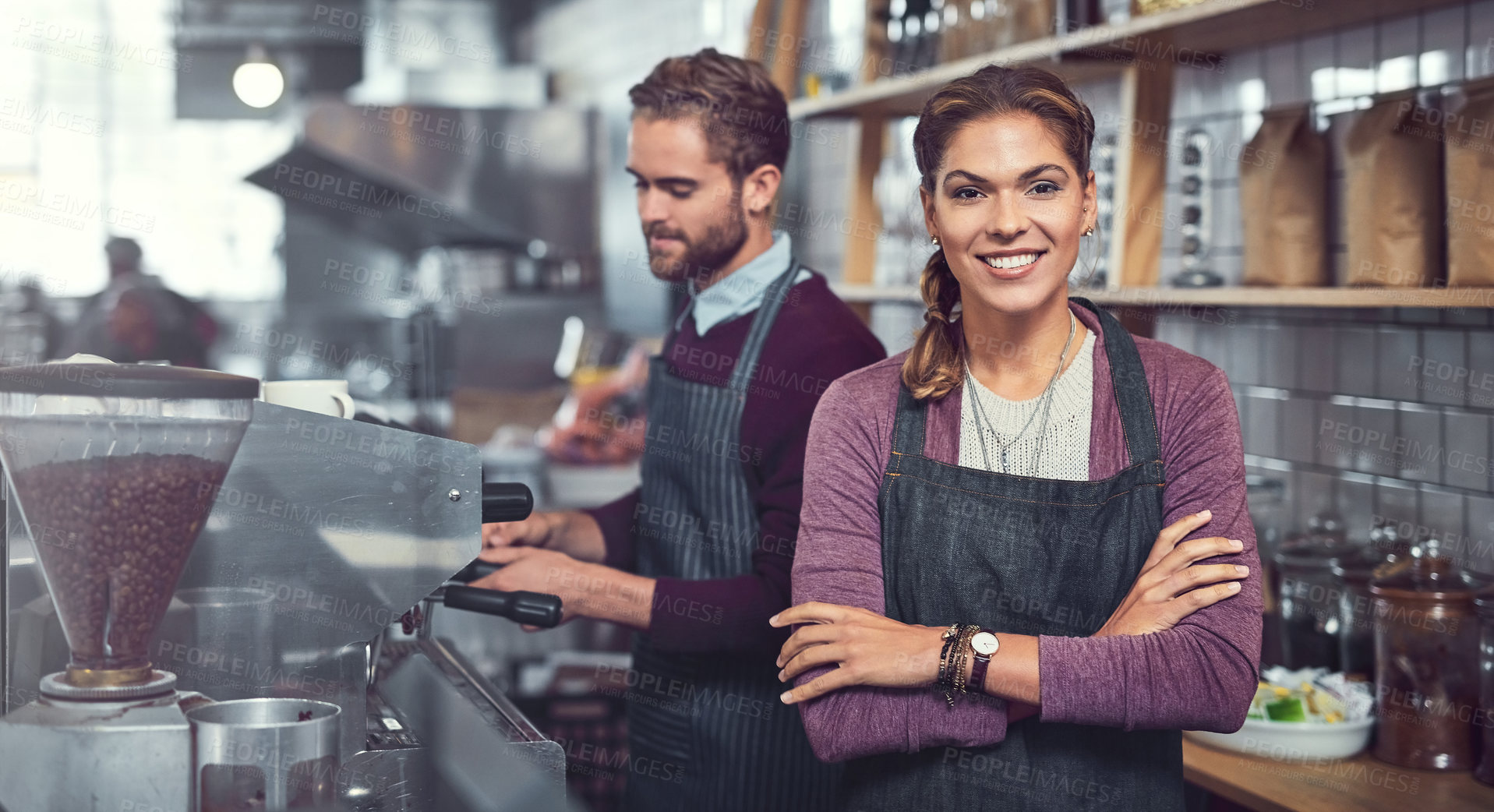 Buy stock photo Portrait of a confident young woman working in a cafe while her coworker operates a coffee machine in the background