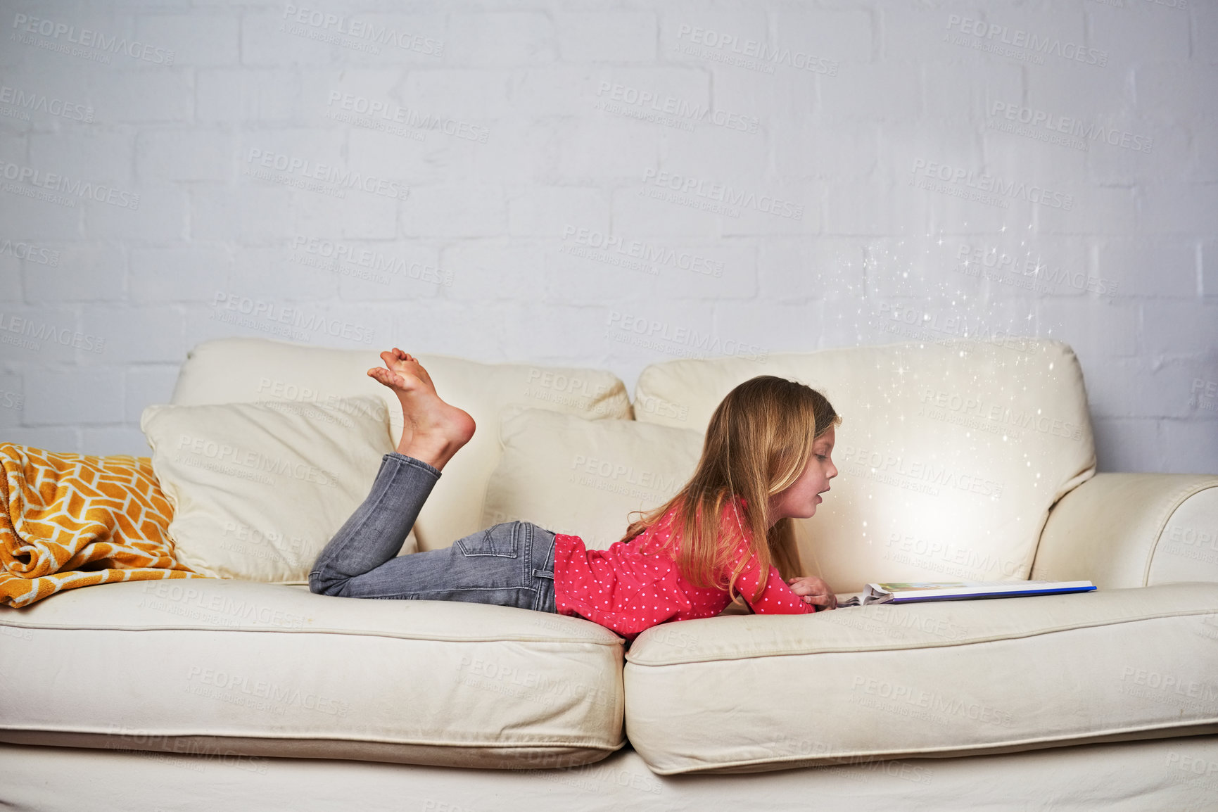Buy stock photo Shot of a little girl reading a book while lying on a sofa