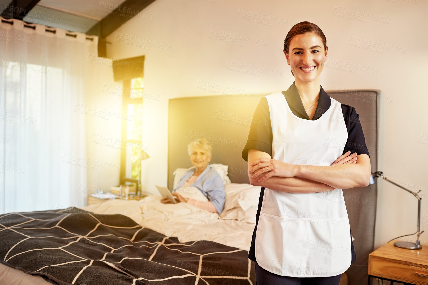 Buy stock photo Portrait of a young nurse standing with her arms folded while her patient sits in bed in the background