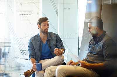 Buy stock photo Shot of two young businessmen talking together while sitting inside a glass office