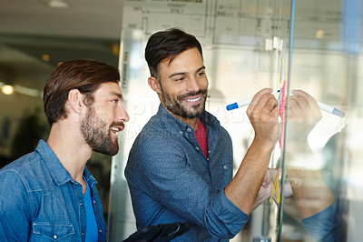 Buy stock photo Shot of two young businessmen brainstorming on a glass wall while standing in an office