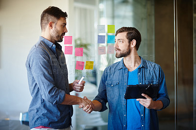 Buy stock photo Shot of two young businessmen shaking hands while brainstorming on a glass wall in an office