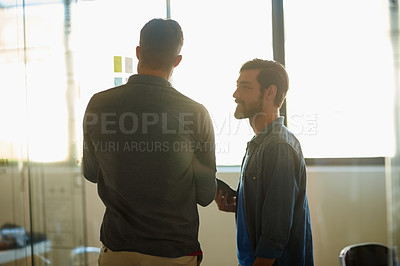 Buy stock photo Shot of two young businessmen brainstorming together while standing inside a glass office