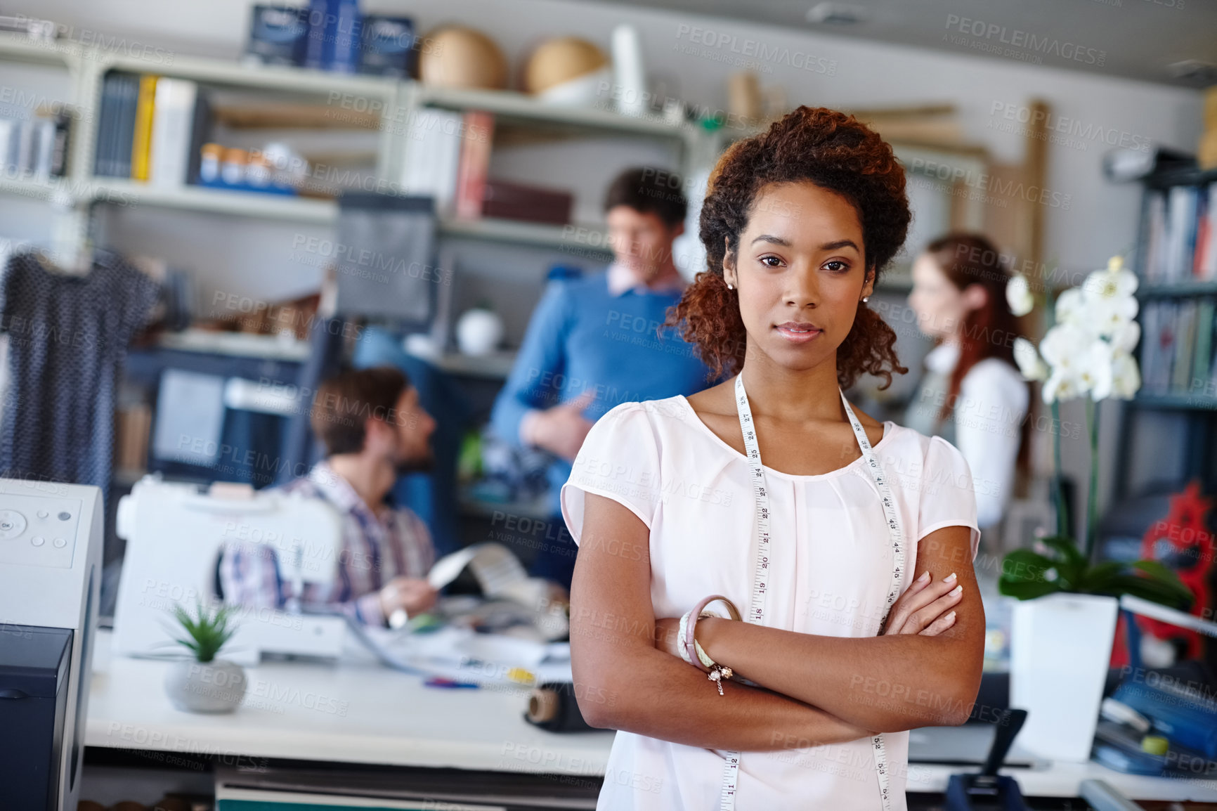 Buy stock photo Portrait of a young fashion designer standing with her arms folded while her colleagues work in the background