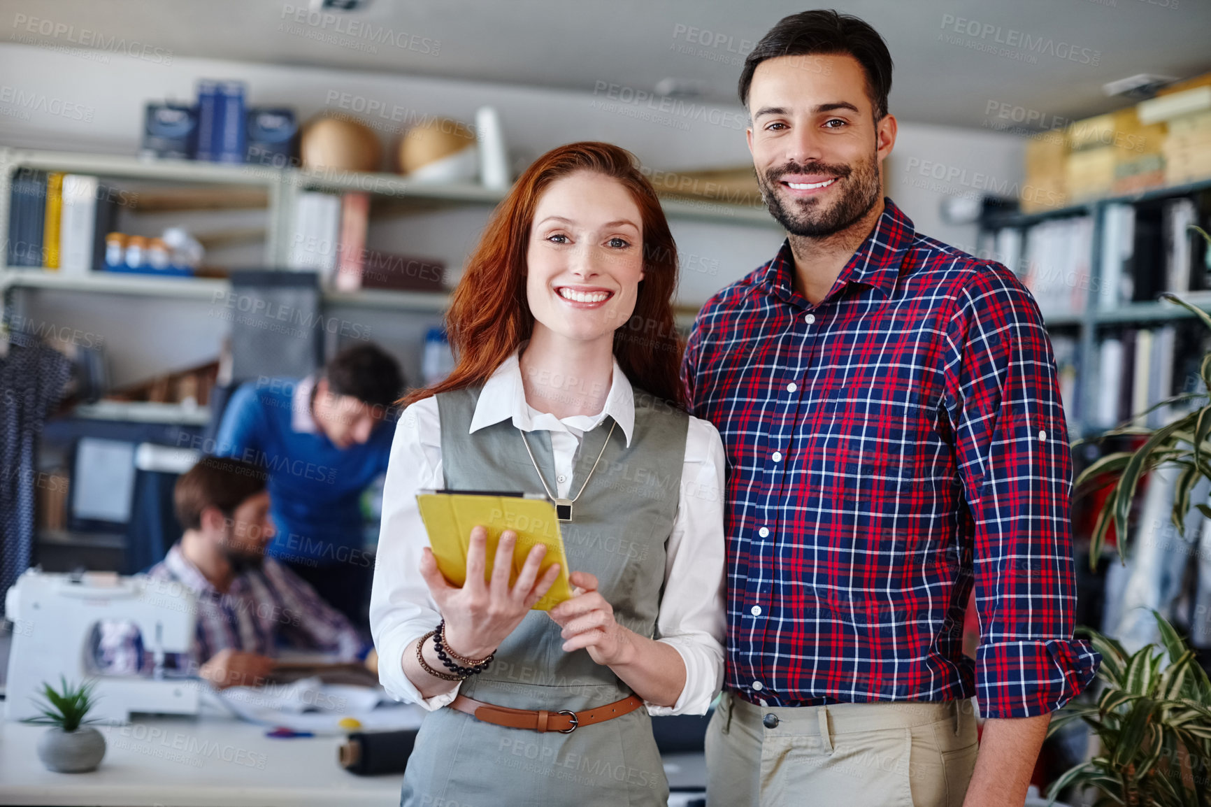 Buy stock photo Portrait of two young fashion designers standing in the workshop while their colleagues work in the background