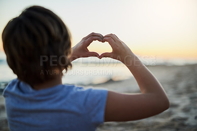 Buy stock photo Rear view shot of a young girl making a heart shape with her hands at the beach