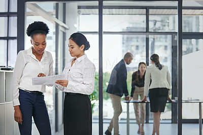 Buy stock photo Shot of two female work colleagues discussing business in the office while their coworkers work in the background