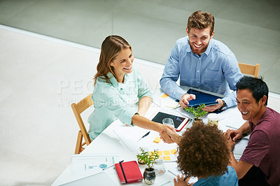 Buy stock photo High angle shot of two businesspeople shaking hands together at a table in an office while colleagues look on