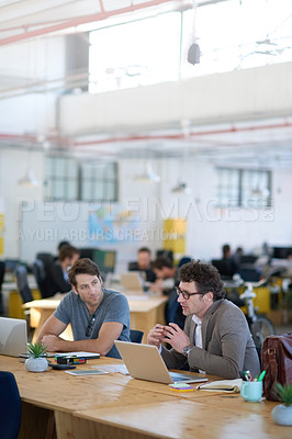 Buy stock photo Shot of two colleagues talking together while working on laptops at a desk in a large office
