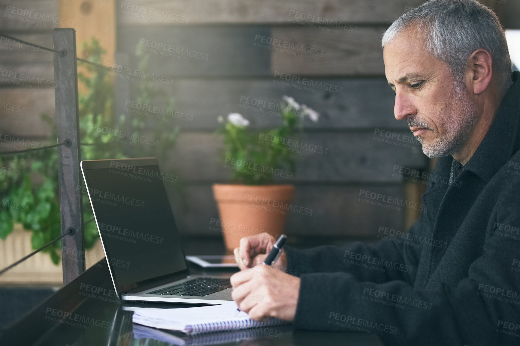 Buy stock photo Shot of a mature businessman writing notes while using his laptop in a cafe