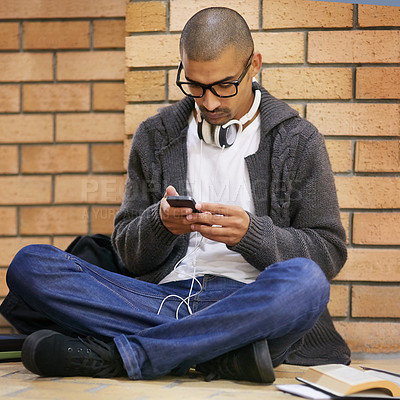 Buy stock photo Shot of a college student texting on his cellphone while sitting in a hallway at campus