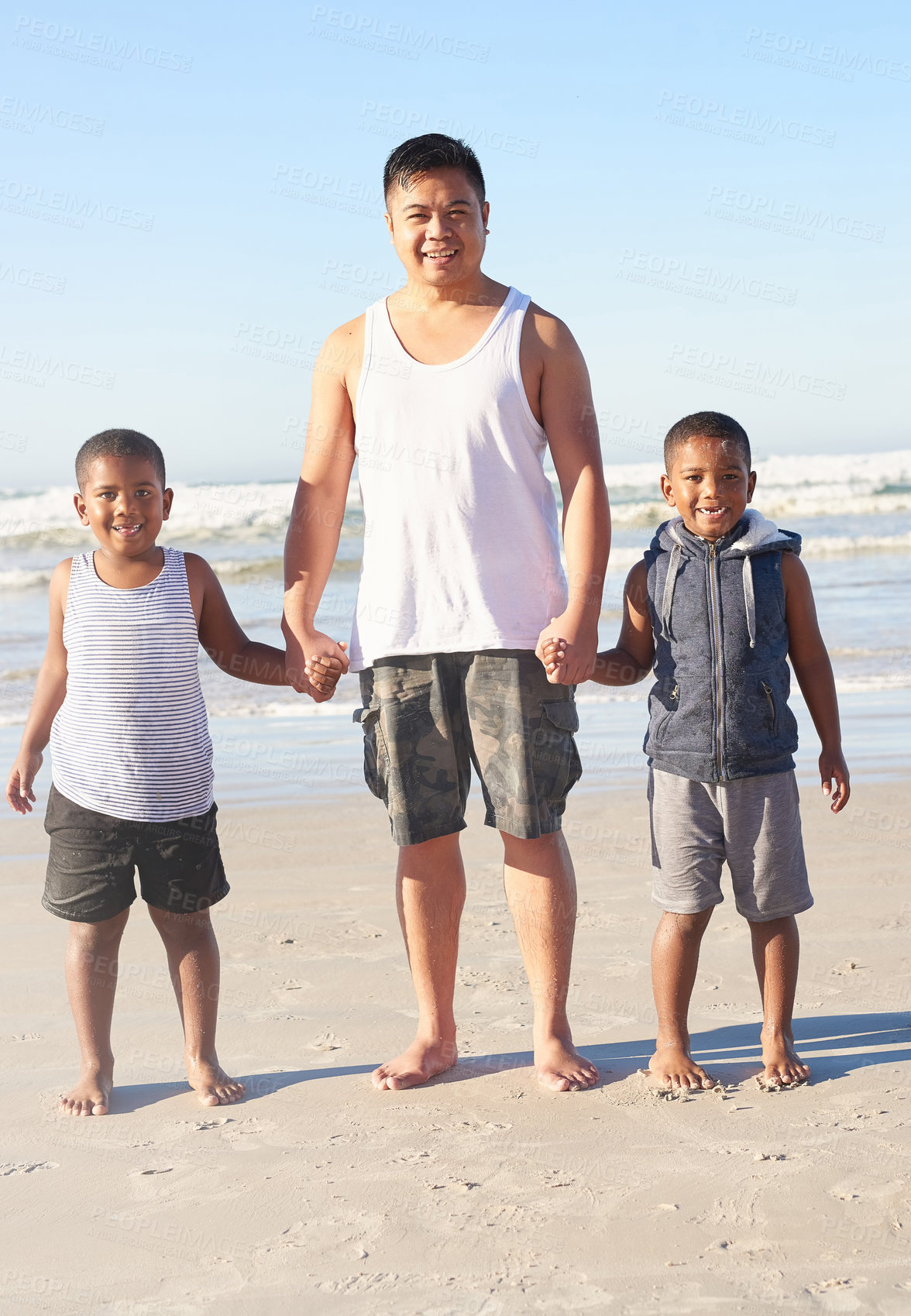 Buy stock photo Portrait of a father and his two sons holding hands while standing on the beach together