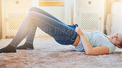 Buy stock photo Woman, home and struggle with jeans on floor for dressing, weight gain, force and tight. Lens flare, female person and bedroom with lying on floor to squeeze small pants for outfit, fashion and style