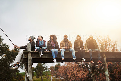 Buy stock photo Shot of a group of friends sitting on a pier together on a weekend breakaway