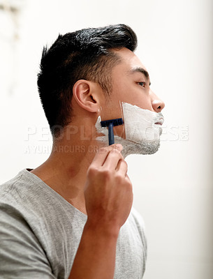 Buy stock photo Shot of a serious young man shaving his beard at home