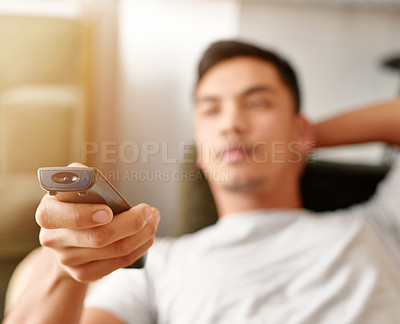 Buy stock photo Shot of a young man using a remote control to change the channel on his television while he lies on the couch