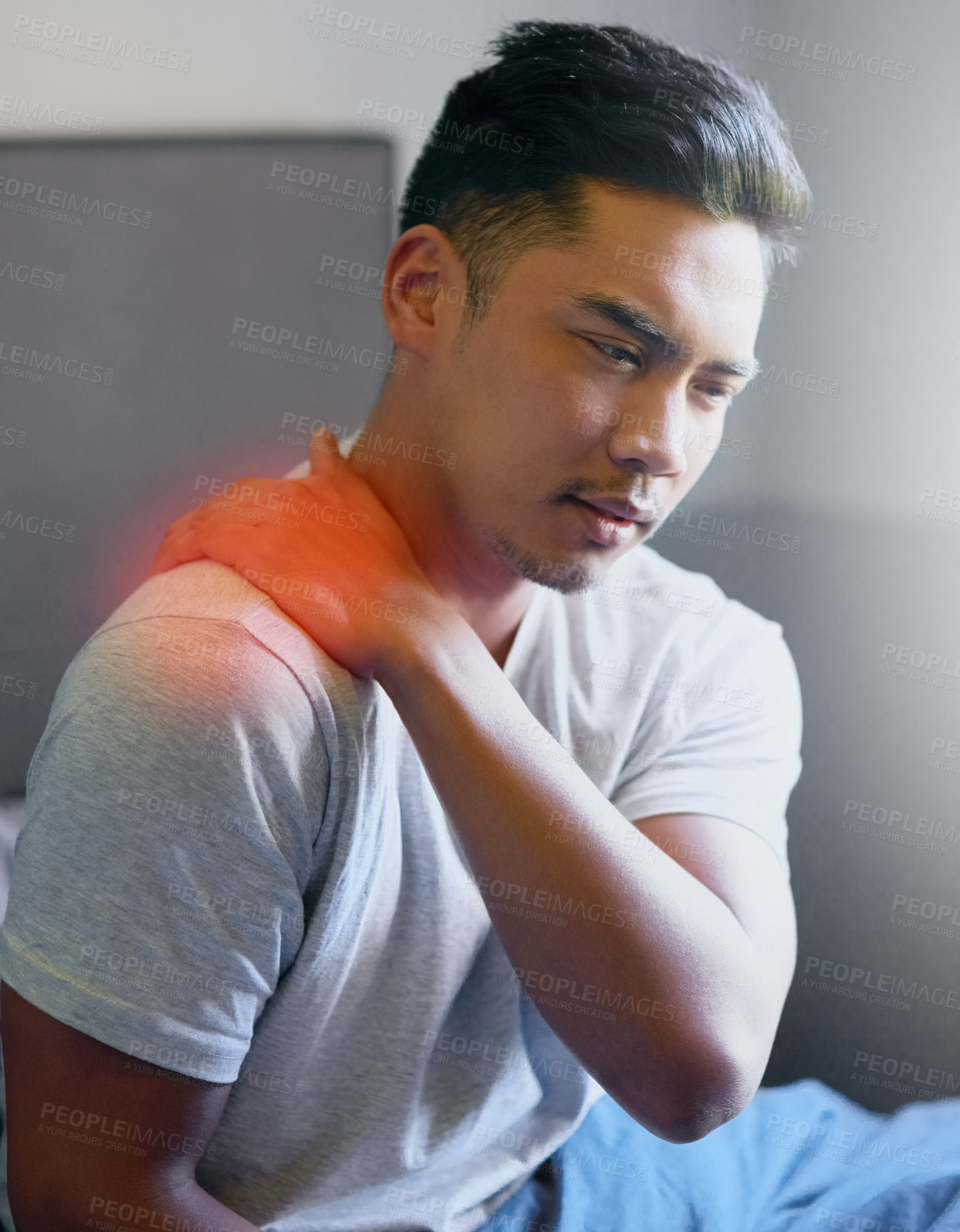 Buy stock photo Shot of a young man rubbing his sore shoulder while sitting up in bed