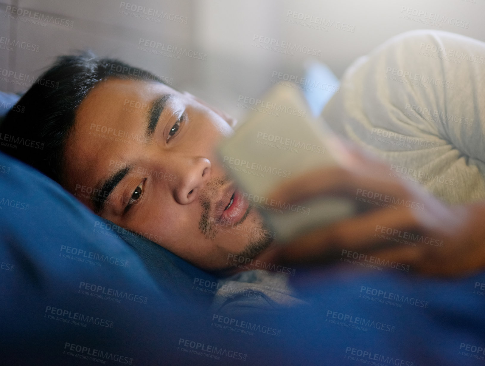 Buy stock photo Shot of a young man looking at his phone while lying in bed