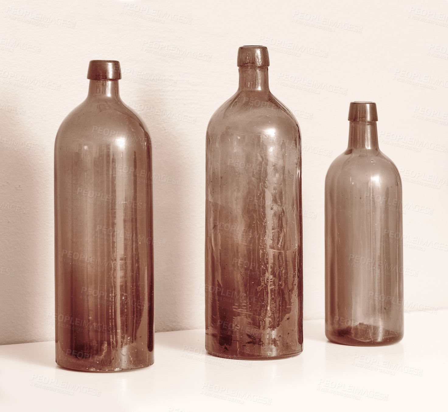 Buy stock photo Old empty glass bottles used for decoration or vases on a table against a white wall. Collection of three brown disposable and transparent vintage containers in different sizes to recycle or reuse
