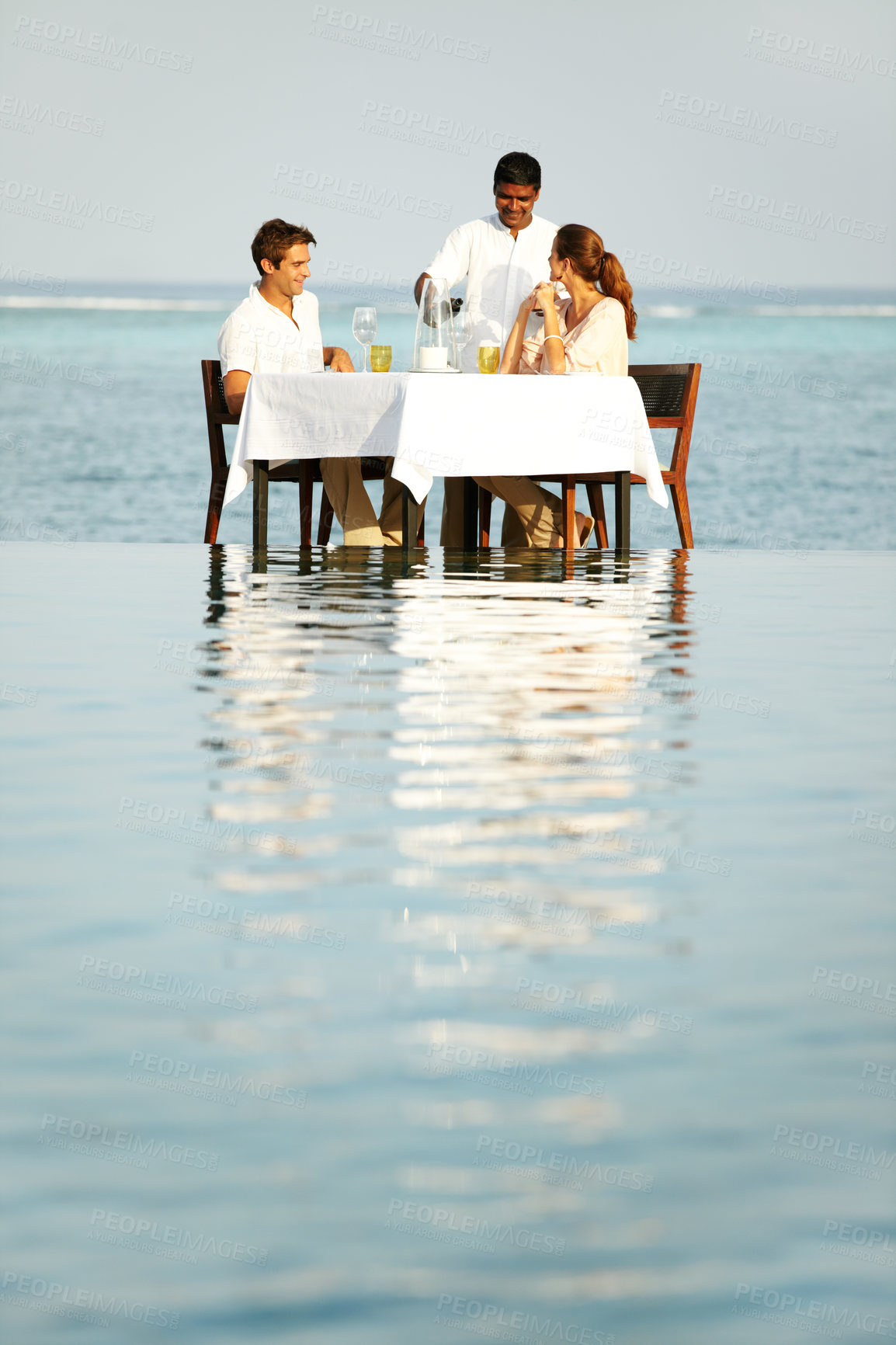 Buy stock photo A young couple sitting at a table outside surrounded by water and being served wine
