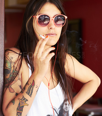 Buy stock photo Shot of a beautiful young tattooed woman smoking a cigarette while listening to music