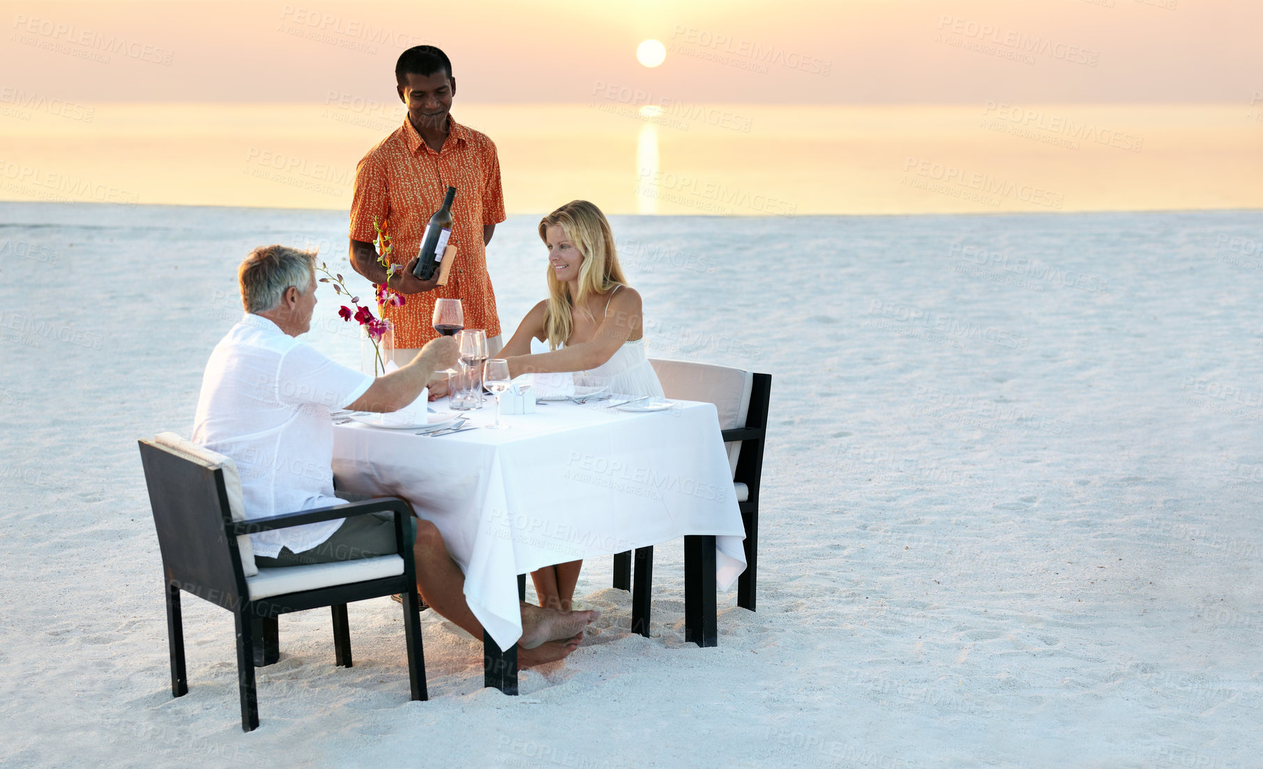 Buy stock photo Couple, beach and dinner in sunset with waiter for romantic outdoor date, valentines day or anniversary. Man and woman with wine, cheers or celebration on table in relax or romance by ocean coast