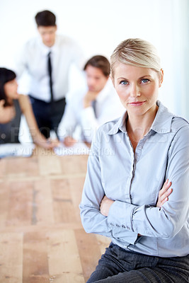 Buy stock photo Portrait of a pretty businesswoman sitting on a desk with her arms folded - blurred colleagues in the background