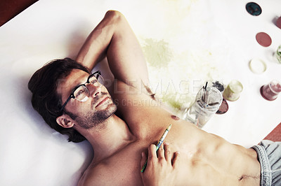 Buy stock photo Artist, lying and thinking with paintbrush for relaxing in space with canvas, paints or jars for work. Young man, face and glasses for inspiration of creative, project or hobby by shirtless in room