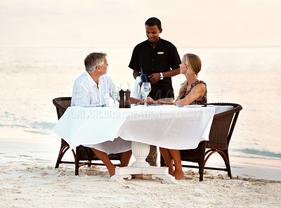 Buy stock photo Beach, dinner and couple with sunset, love and romance with anniversary, waiter and vacation. People, romantic meal or man with woman, ocean and waves with summer holiday, travel or tropical island