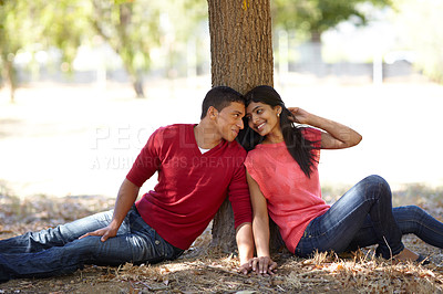 Buy stock photo Shot of an affectionate young couple sitting together under a tree in the park